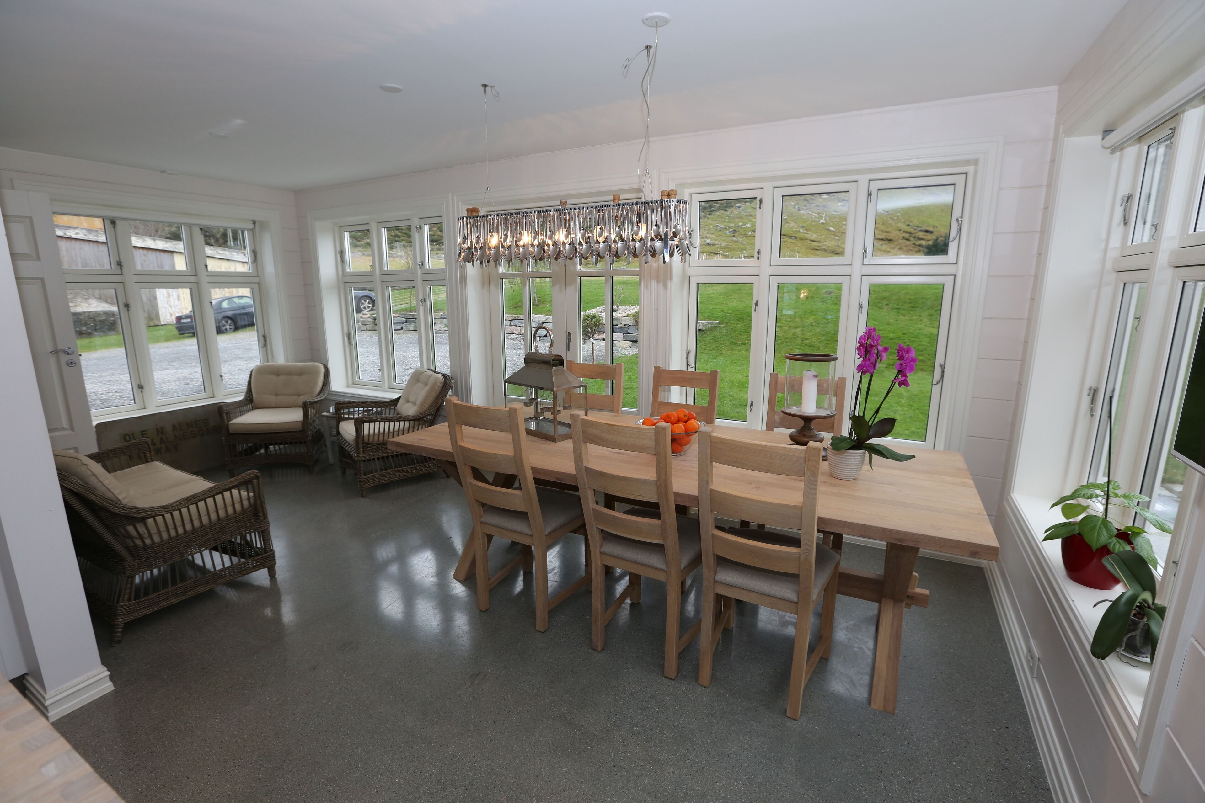Un comedor con mesa y sillas de madera, una araña y ventanas amplias con vistas al exterior.