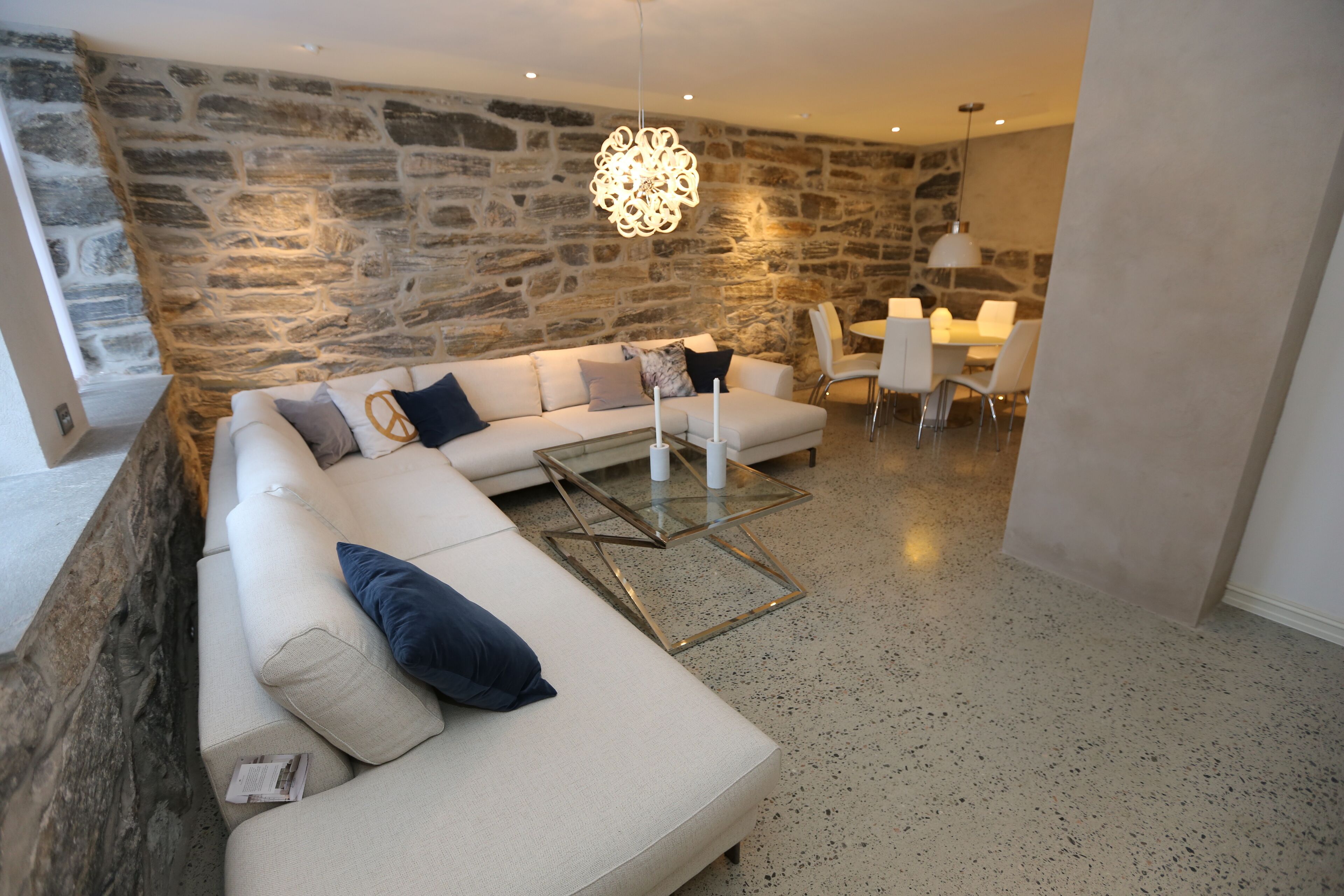 Un salon moderne avec un mur en pierre, un canapé blanc agrémenté de coussins bleus, une table basse en verre et un coin repas avec une table et des chaises blanches.
