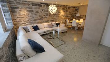 Un salon moderne avec un mur en pierre, un canapé blanc agrémenté de coussins bleus, une table basse en verre et un coin repas avec une table et des chaises blanches.