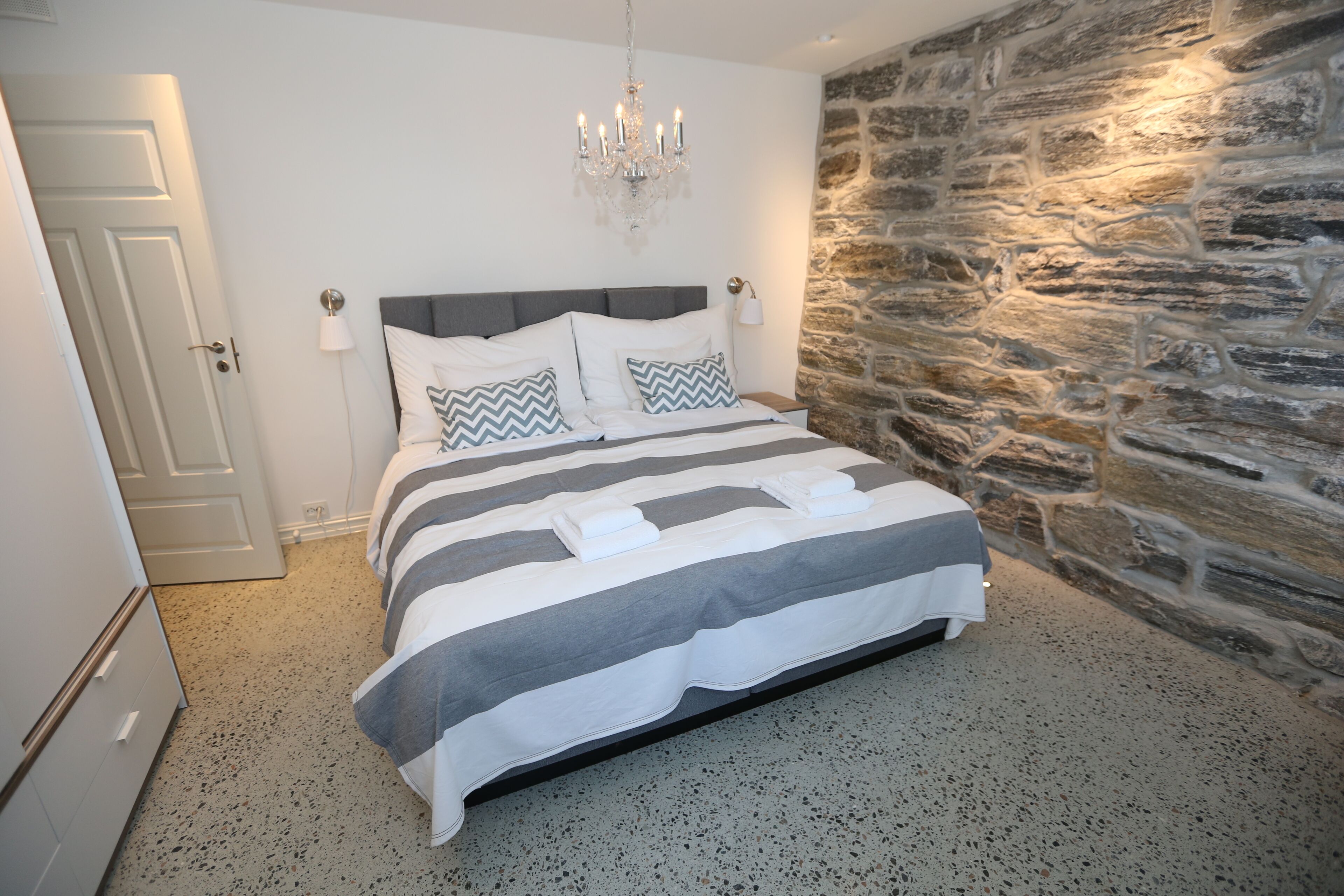 A bedroom with a bed, a chandelier, a stone wall, and a door.