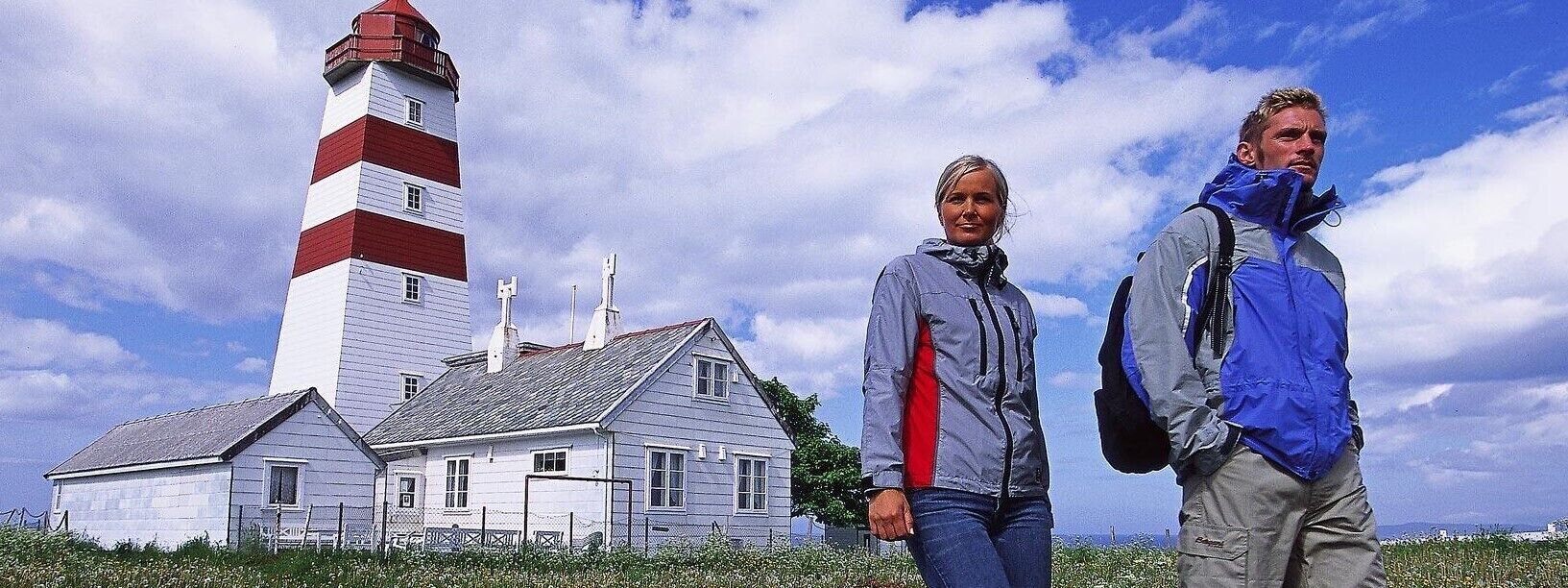 A lighthouse with a white and red striped tower, a white building with a grey roof, and a clear blue sky with clouds.
