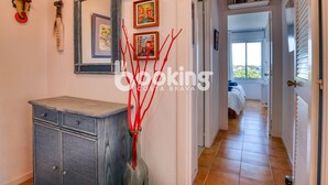 Un pasillo con un aparador antiguo, obras de arte enmarcadas y un jarrón con ramas rojas. Hay puertas que dan a un dormitorio con vistas al exterior.