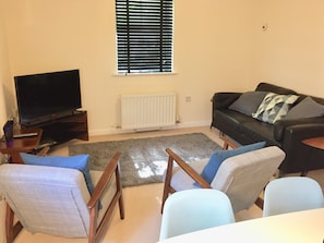A living room with a flat-screen TV, a black leather sofa, two armchairs, a grey rug, and a window with blinds.