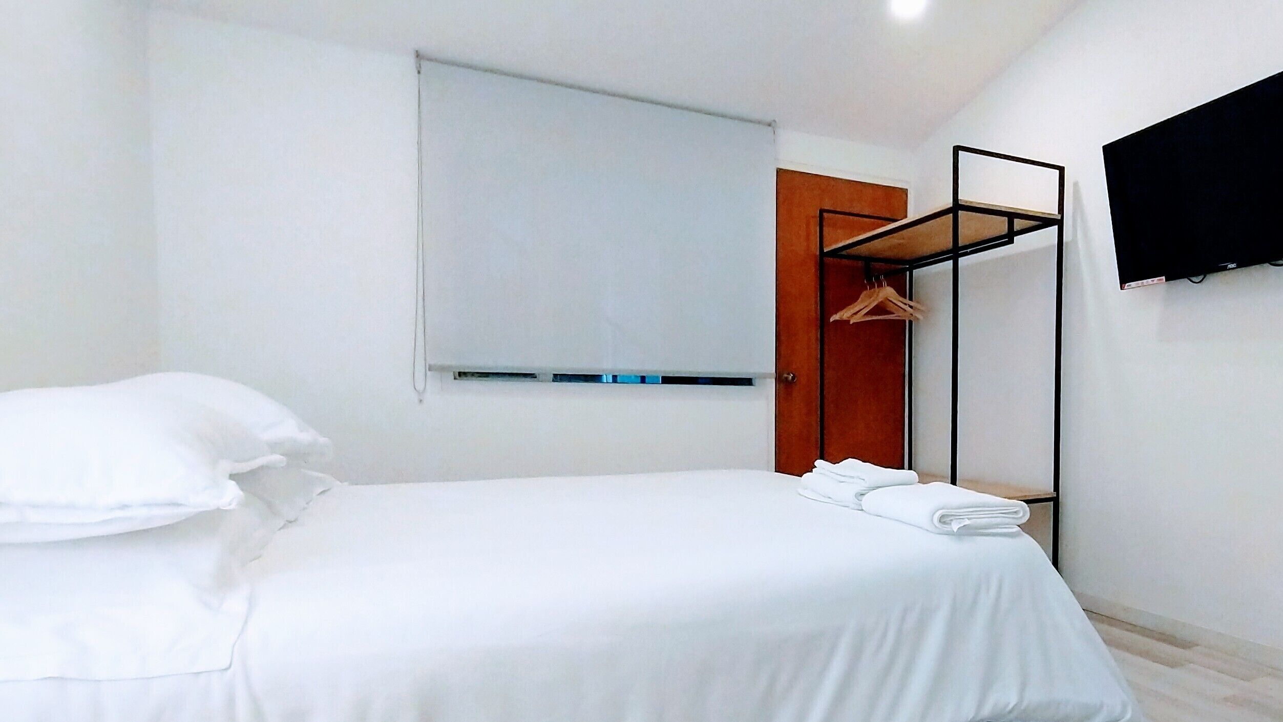 A hotel room with a bed, a wooden door, a coat rack, and a flat-screen TV mounted on the wall.