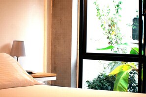 A bedroom with a bed, a bedside table with a lamp, and a window with a view of greenery.