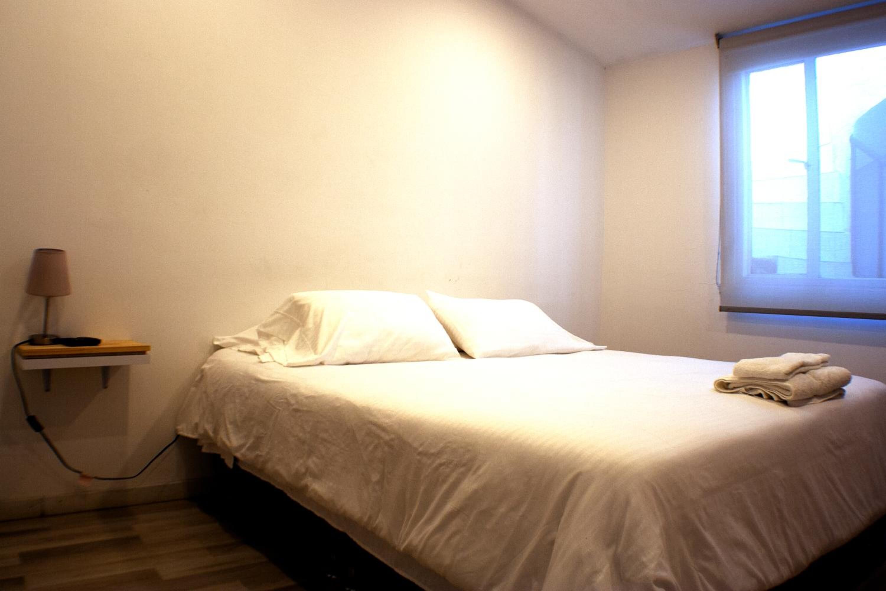 Cama bem arrumada com lençóis brancos, abajur e janela com persianas.