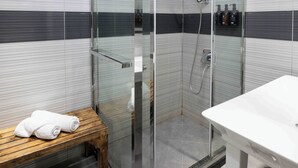 A modern bathroom with a glass-enclosed shower, a wooden bench with towels, and a white sink. - Sleepover Terminal 1, Concourse D – formerly sleep ‘n fly (Dubai)