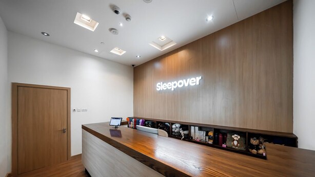 A reception desk with a 'Sleepover' sign, a computer monitor, and a shelf with various items. - Sleepover Terminal 1, Concourse D – formerly sleep ‘n fly (Dubai)