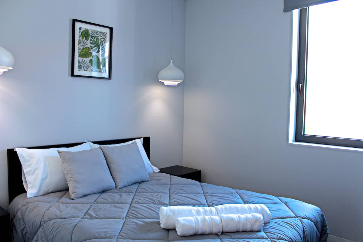 A neatly made bed with a grey duvet and pillows, a framed tropical leaf artwork on the wall, and a window with daylight coming through.