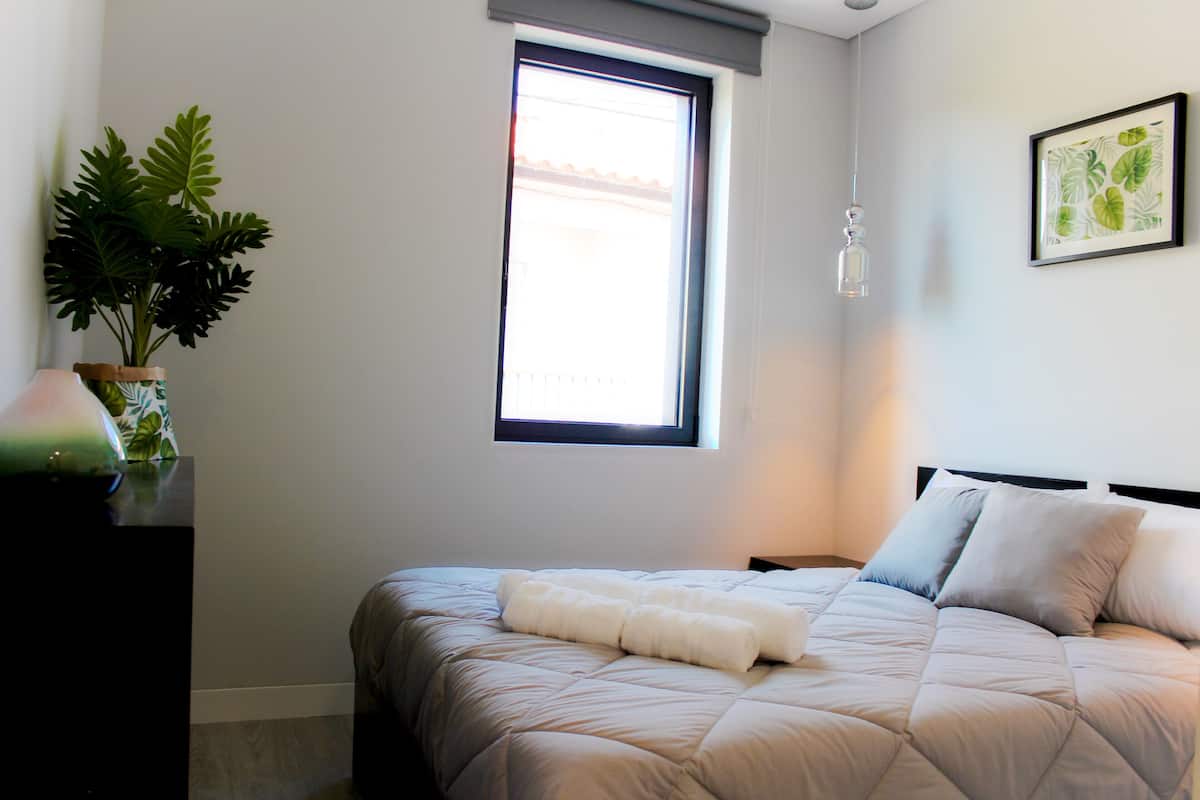 A bedroom with a bed, a window, a plant, and a framed picture on the wall.