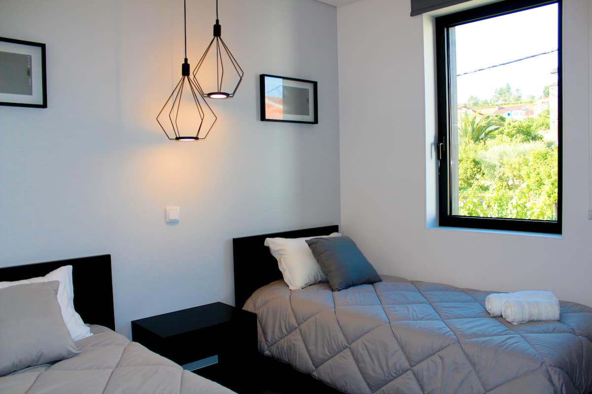 A bedroom with two beds, a window with a view of greenery, and pendant lights.