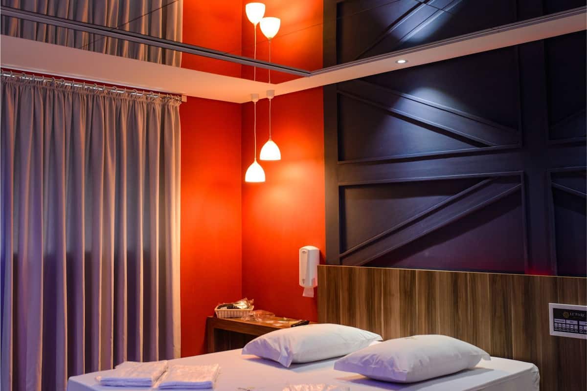 A hotel room with a bed, curtains, a wooden panel wall, and a small table with a phone and a basket.