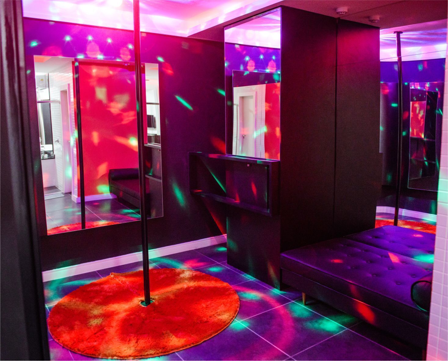 Quarto com tapete vermelho, sofá preto e parede espelhada refletindo uma porta e um corredor.
