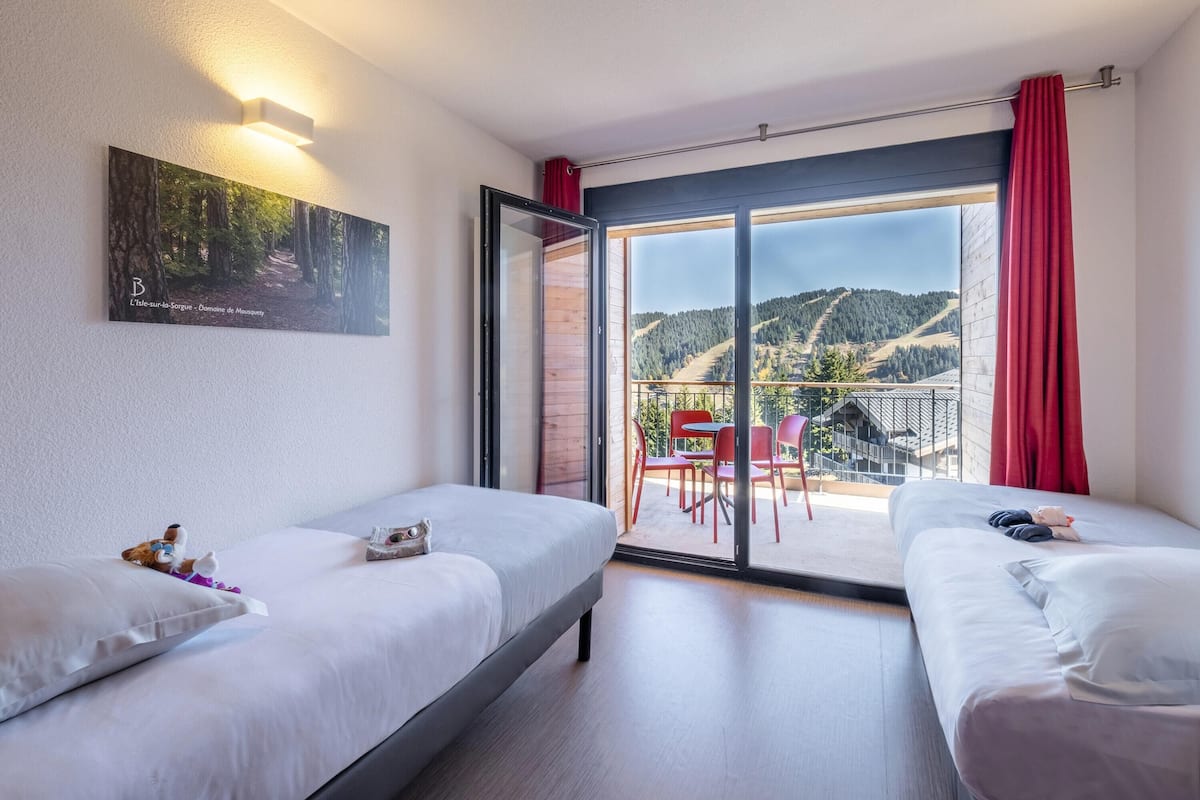 Ein Hotelzimmer mit zwei Betten, einem großen Fenster mit Blick auf Wald und Berge sowie einem Balkon mit roten Stühlen.