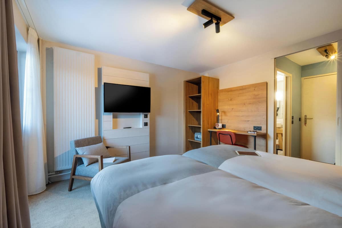 Ein Hotelzimmer mit zwei Betten, einem an der Wand befestigten Fernseher, einem Holztisch und einem Kleiderschrank.