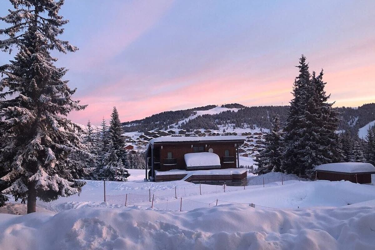 Eine Holzhütte, umgeben von schneebedeckten Bäumen und einem Bergpanorama.