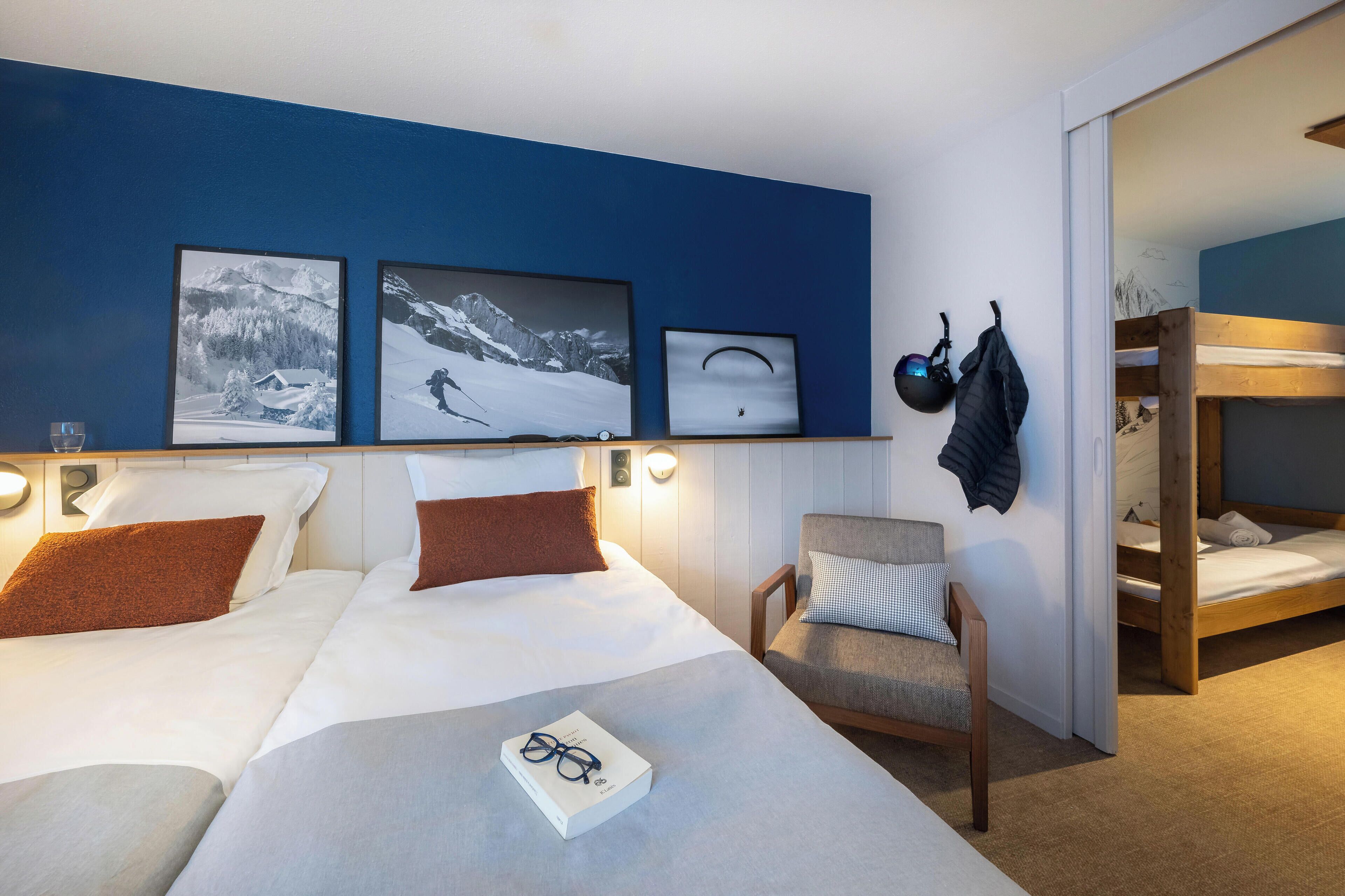 Une chambre d’hôtel avec deux lits, une chaise et un lit superposé. Des tableaux encadrés ornent le mur, et un casque est accroché au mur.