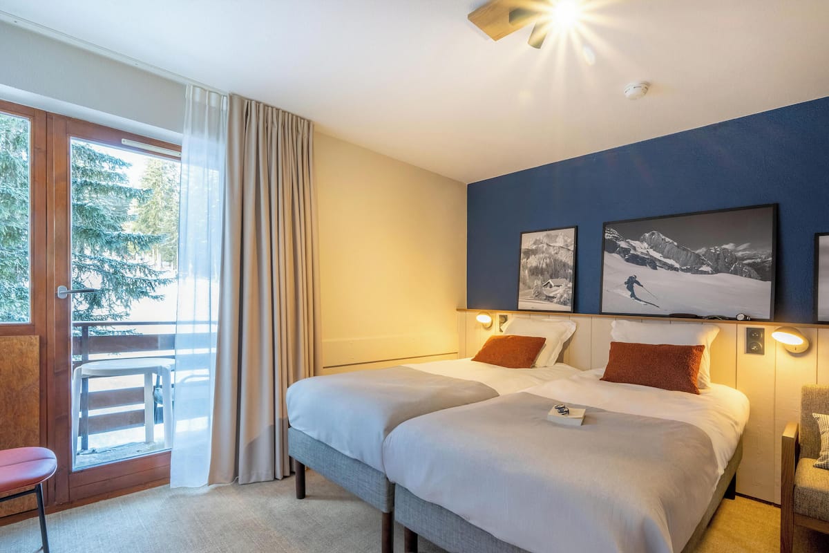Ein Hotelzimmer mit einem großen Bett, zwei Nachttischen, einem Sessel, einem Balkon mit Blick auf Bäume und zwei gerahmten Bildern an der Wand.