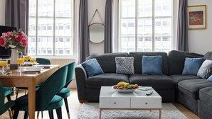 A modern living room with a grey sofa, teal chairs, a coffee table with fruit, and a dining area with a table set for two. - 3-bedroom apartment Lancaster gate (London)