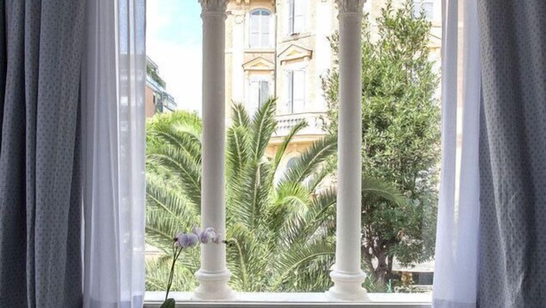 Habitación con ventana que da a un edificio con arcos, una pequeña mesa con una planta y un banco.