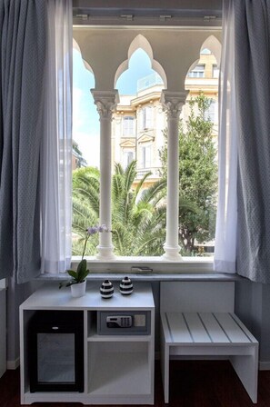 Habitación con ventana que da a un edificio con arcos, una pequeña mesa con una planta y un banco.