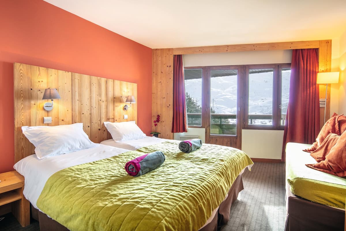 A hotel room with two beds, wooden headboard, large windows with curtains, and a view of the mountains.