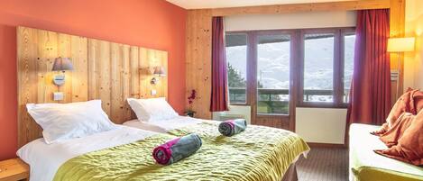 Ein Hotelzimmer mit zwei Betten, einem Holzkopfstück, großen Fenstern mit Vorhängen und Bergblick.