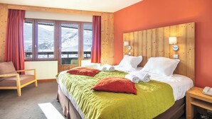 Habitación de hotel con una cama grande, cabecero de madera, dos lámparas de noche, una silla y vistas a las montañas desde las ventanas.