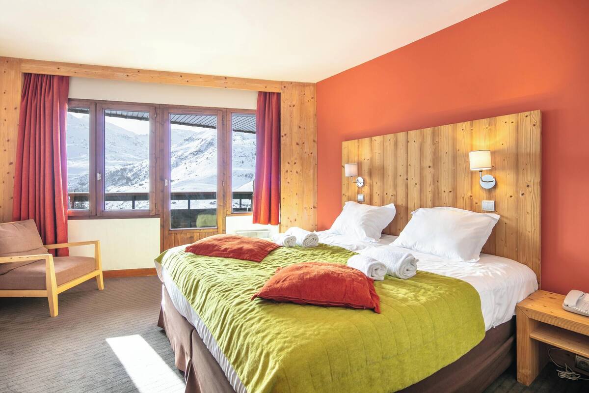 A hotel room with a large bed, wooden headboard, two bedside lamps, a chair, and a view of mountains through the windows.