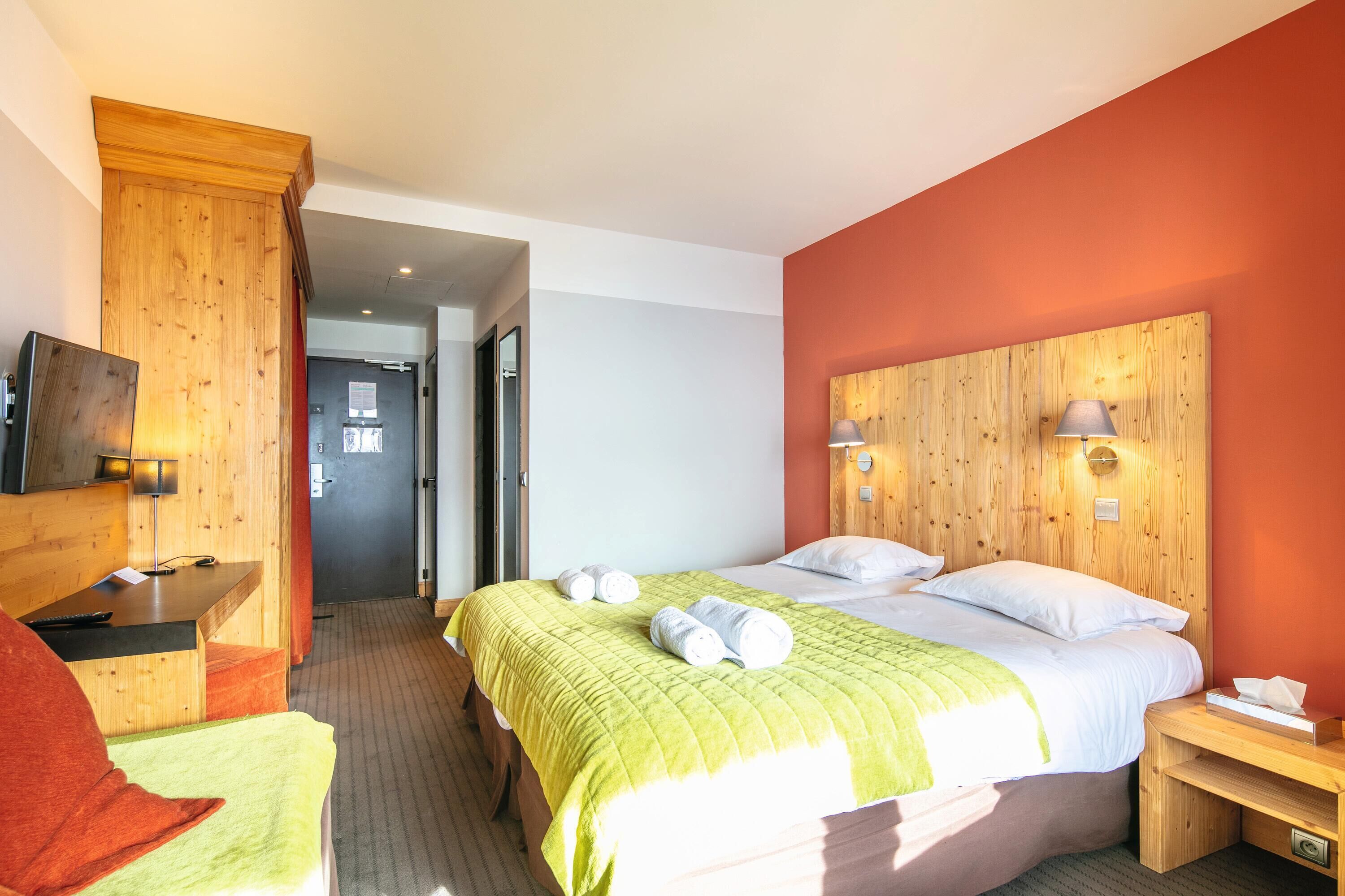 Une chambre d’hôtel avec une tête de lit en bois, un lit avec une literie verte, deux lampes de chevet et une table de chevet en bois avec une lampe.