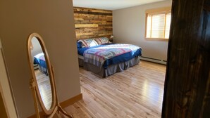 A bedroom with a wooden headboard, a large bed with a colorful blanket, a wooden floor, and a window with blinds.