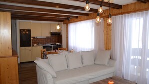 A cozy living room with a sofa, wooden coffee table, and pendant lights. A kitchen is visible in the background with wooden cabinets and a dining area. - Nice flat for rent in Les Collons (Thyon-Les Collons)