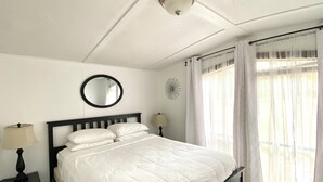 Een netjes opgemaakt bed met een witte dekbedovertrek, een ronde spiegel aan de muur en een raam met dunne gordijnen.