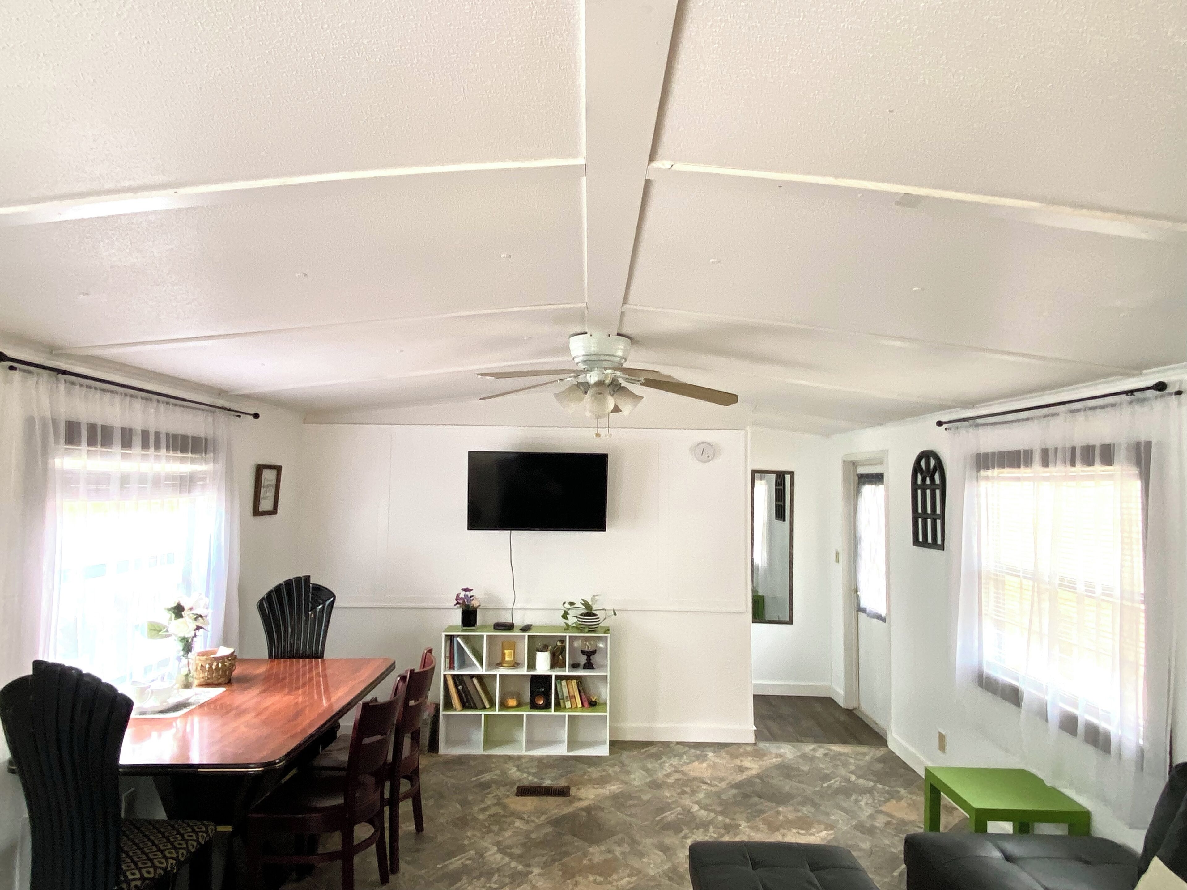 A dining area with a wooden table and chairs, a ceiling fan, a TV mounted on the wall, and a bookshelf with various items.