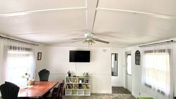 A dining area with a wooden table and chairs, a ceiling fan, a TV mounted on the wall, and a bookshelf with various items.