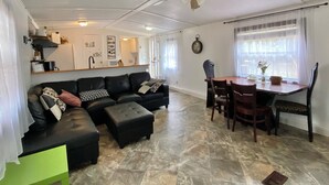 Een woonkamer met een bank, een poef, een eettafel met stoelen, een plafondventilator en een klok aan de muur.