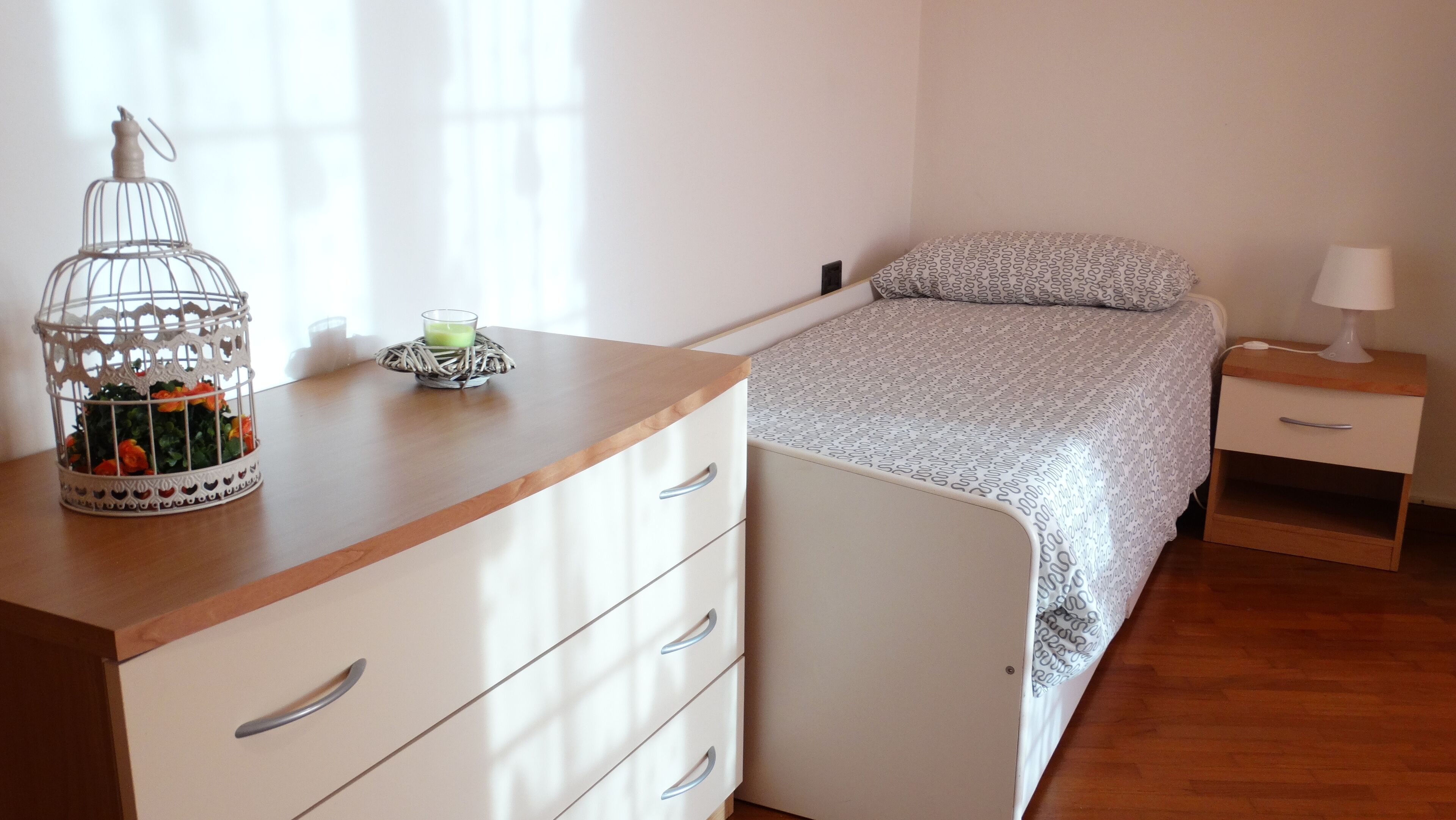 Un dormitorio con cama, mesita de noche, cómoda, lámpara y una jaula decorativa con flores.