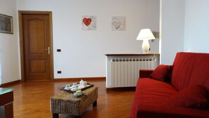 A living room with a wooden door, a red sofa, a wicker ottoman, and a radiator. - ..... SUPERCASONA PRATICA! (Sarzana)