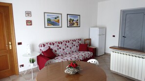 Una sala de estar acogedora con un sofá, una mesa de comedor con un centro de mesa, un refrigerador y dos cuadros en la pared.