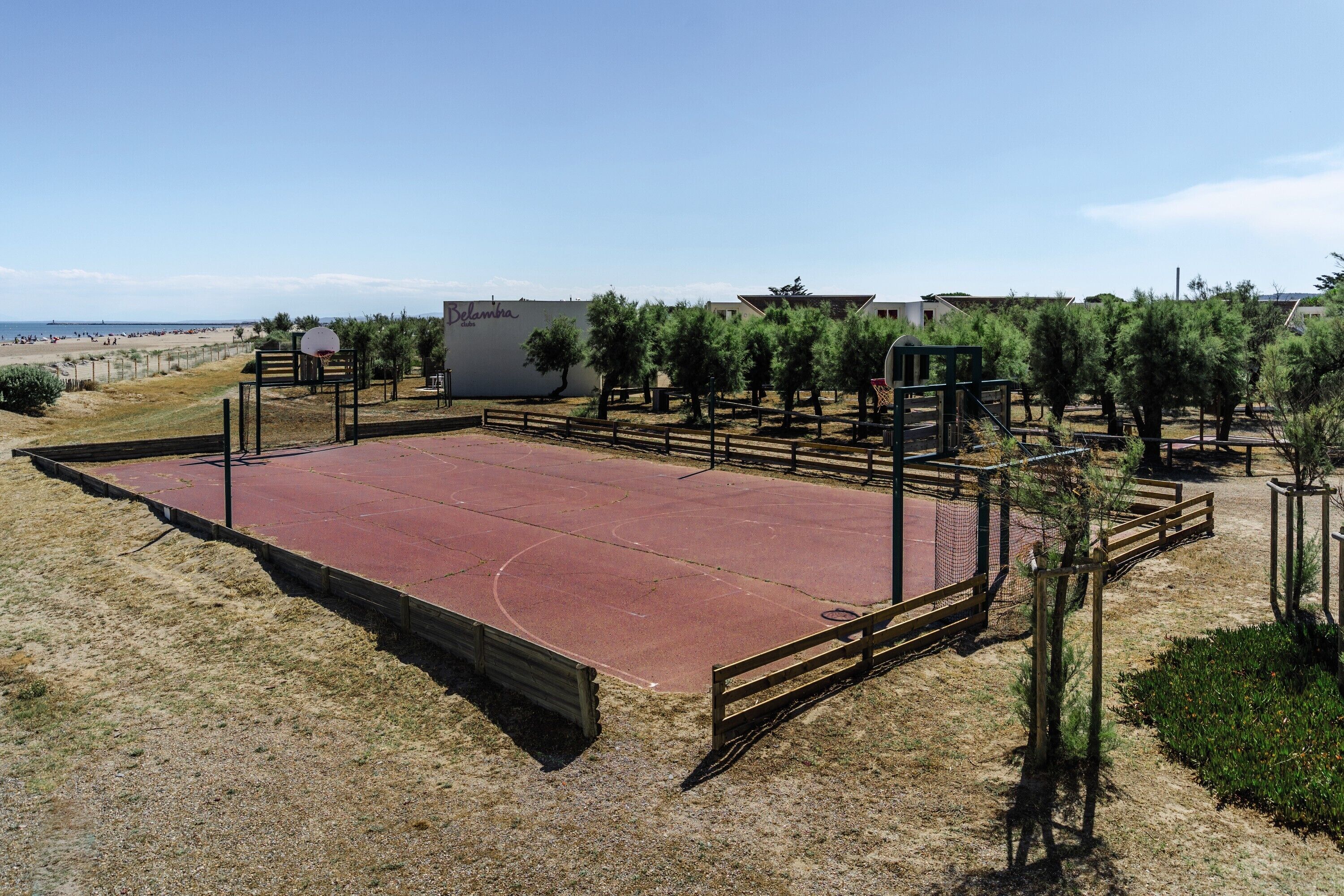 Ein Basketballplatz im Freien mit roter Oberfläche, umgeben von Zäunen und Bäumen, mit Strand und Gebäuden im Hintergrund.