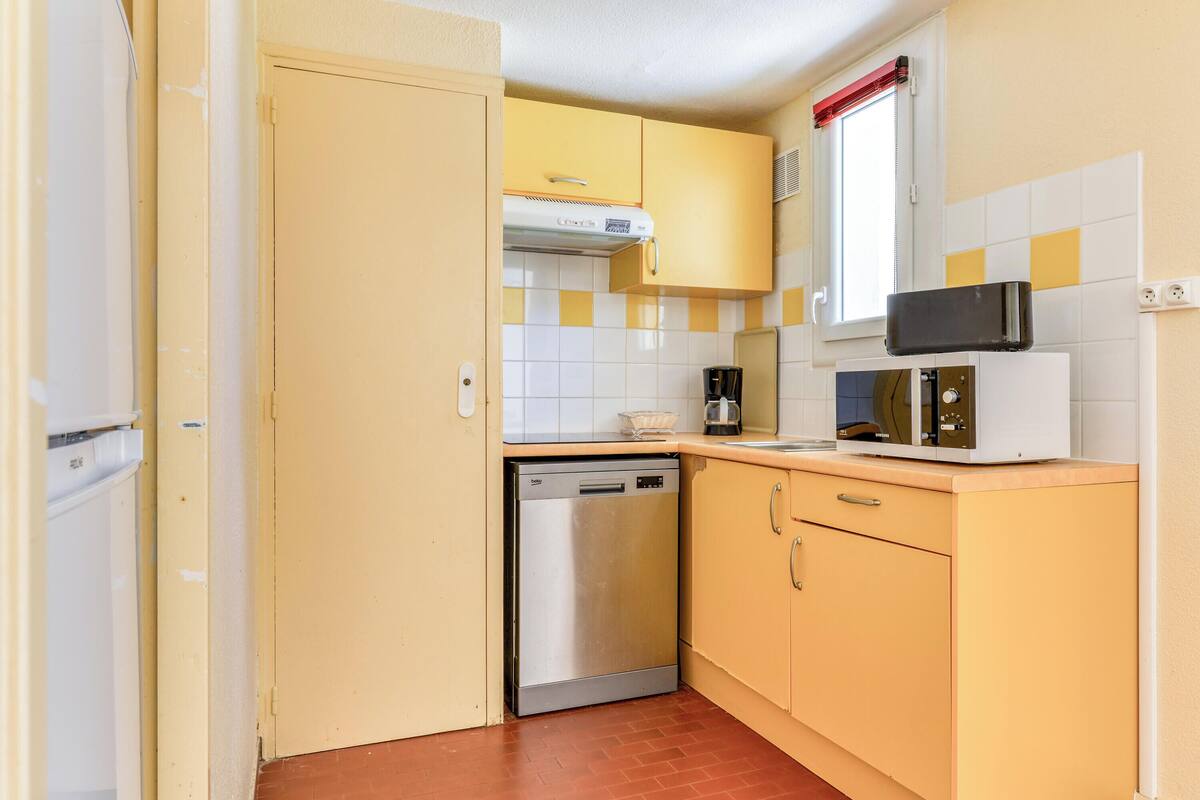 A compact kitchen with yellow cabinets, a stainless steel dishwasher, a microwave, and a window with a red blind.