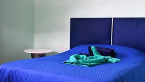 A bed with a blue mattress and a green blanket, a round table with a pink top, and a white wall with a power outlet.