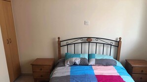 Een bed met een quilt in blauw, roze en grijs, twee houten nachtkastjes en een houten garderobe.