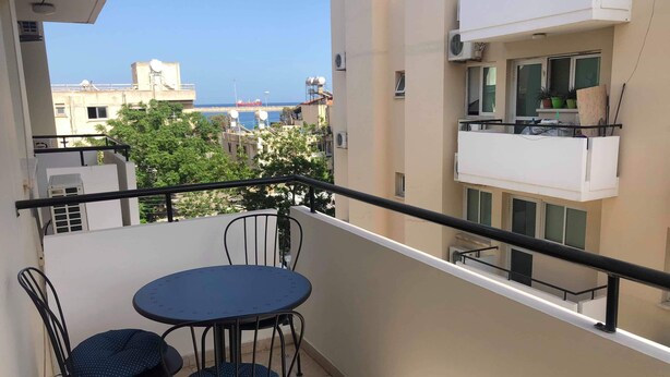 Een balkon met een ronde tafel en twee stoelen, met uitzicht op een stadsgezicht met gebouwen en een schip in de verte.