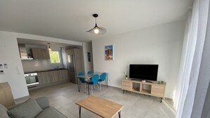 A modern living room with a grey sofa, a wooden coffee table, and a TV on a wooden stand. There is a dining area with blue chairs and a kitchen in the background. - Calvi bright T3 top floor 65m2 panoramic view, 4 people, 2 bedrooms, parking (Calvi)