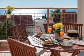 A wooden table with breakfast items, a glass of orange juice, and a view of the sea.