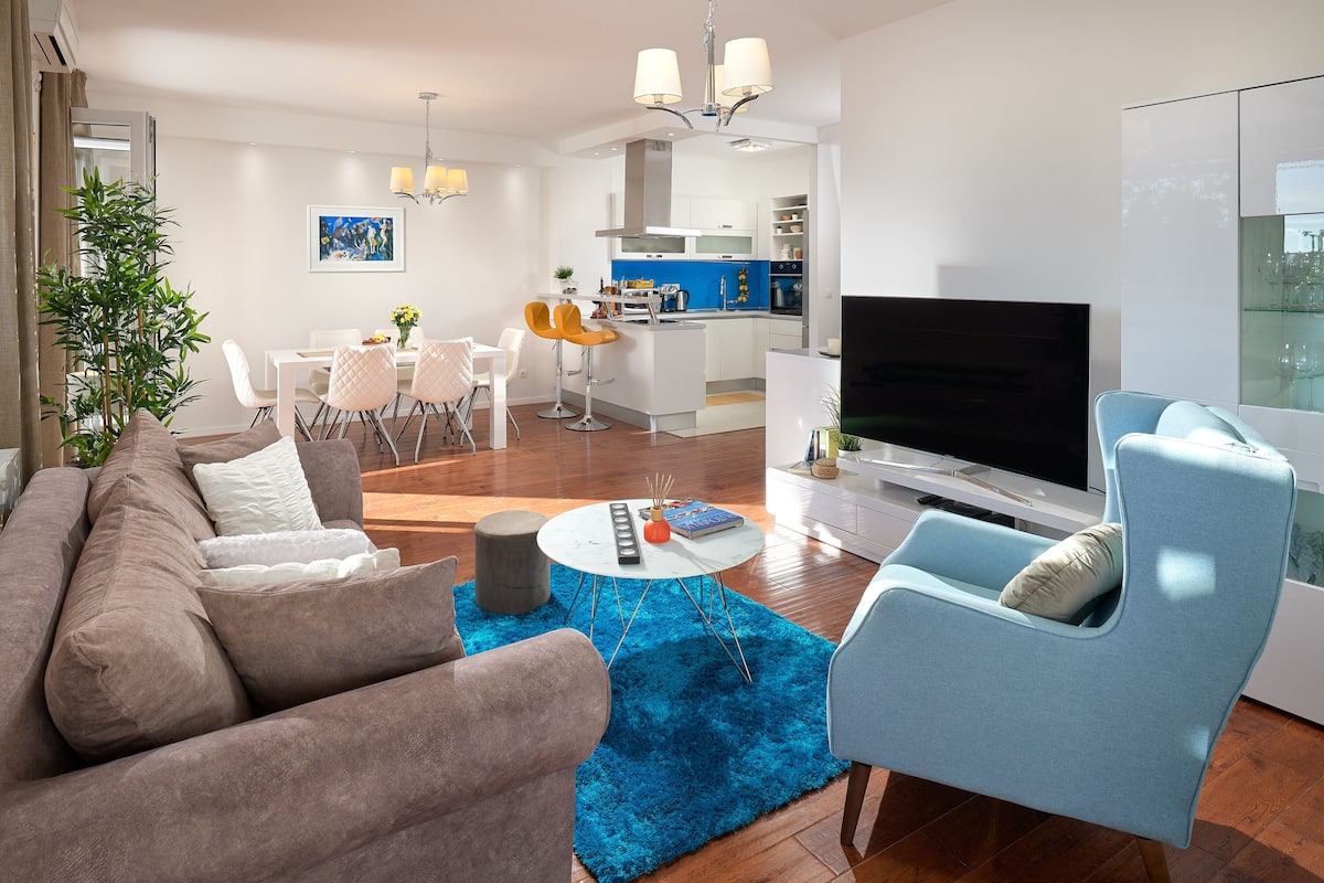 A modern living room with a sofa, a blue rug, a TV, and a kitchen area in the background.