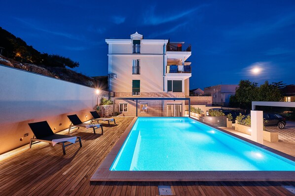 A modern building with a swimming pool, lounge chairs, and a wooden deck.