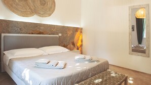A neatly made bed with white linens and two folded towels, a woven wall hanging, a textured wall panel, and a mirror with a wooden frame. - Dimora il Mandorlo -The Plum tree private jacuzzi (Ostuni)