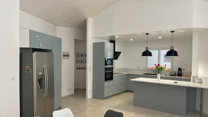 Eine moderne Küche mit grauen Schränken, einem Edelstahlkühlschrank, einem eingebauten Ofen und einer großen Kücheninsel mit Spüle. Über der Insel hängen Pendelleuchten und auf der Arbeitsplatte steht eine Vase mit Blumen.